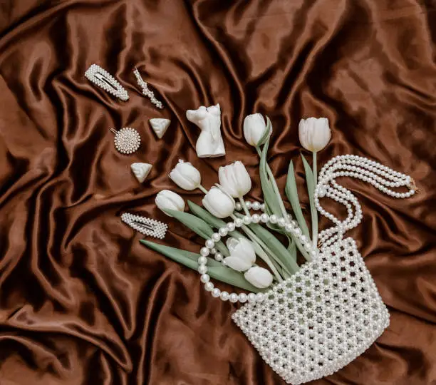 Photo of Flat lay  fashion composition with white tulips, pearl bag, perfume bottle and women's accessories and bijouterie on brown silky background.  lifestyle concept.