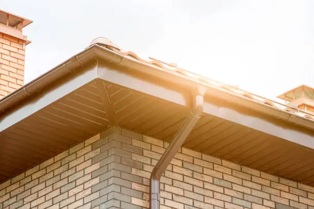 Photo of Corner of house with new brown metal tile roof and rain gutter. Metallic Guttering System, Guttering and Drainage Pipe Exterior