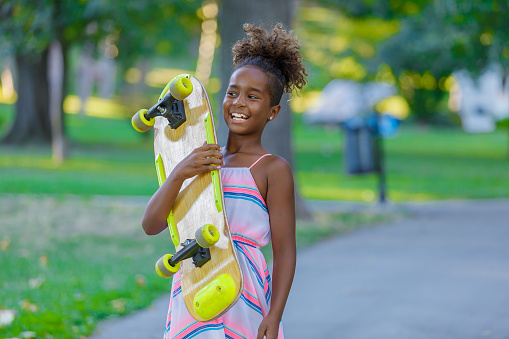 Cute African Girl with Curly Hair, Wearing Dress is Enjoying in Summer Day and Walking With a Long Board in a Path Through a Park and Smiling.