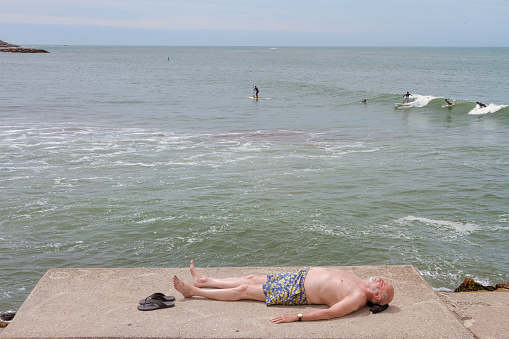 Mar del Plata, Argentina - January 15, 2017: Adult man lying on a coast stone sunbathing while young people surf in the sea of Mar del Plata, Argentina