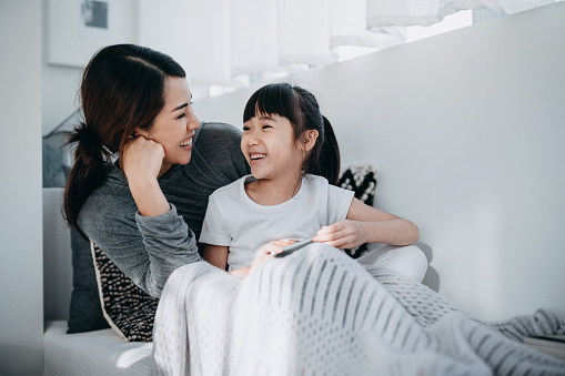 Young Asian mother sitting by the window in the bedroom reading book to little daughter, enjoying family bonding time together at home