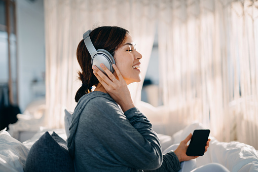 Relaxed young Asian woman with eyes closed sitting on her bed enjoying music over headphones from smartphone at home