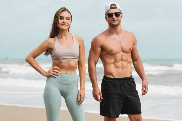 해변에서 함께 서있는 쾌활한 남성과 여성 운동 선수의 초상화. 건강한 남성과 여성은 섹시한 포즈를 취하고 여름날에 근육을 과시합니다. - body women beauty slim 뉴스 사진 이미지