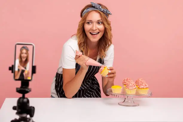 Portrait of happy excited food blogger broadcasting livestream, showing to camera of smart phone process of decorating cakes, using pastry cone. Indoor studio shot isolated on pink background.