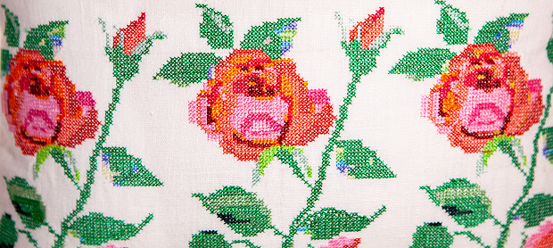 Embroidery in the form of roses on a white pillow that lies on an armchair with a red and white blanket, plants in the background. The interior of a cozy room.
