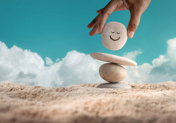 Enjoying Life Concept. Harmony and Positive Mind. Hand Setting Natural Pebble Stone with Smiling Face Cartoon to Balance on Beach Sand stock photo