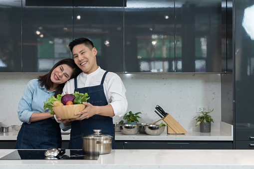 Portrait of a lovely couple in an apron holding a vegetable basket to cooking food in a home kitchen. Asian couple uses free time to cook together in weekends on kitchen island. Culinary lifestyle