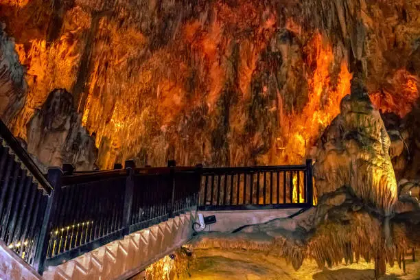 Wooden railing of a staircase against the background of orange-yellow fiery stone formations underground in Damlatas cave (Alanya, Turkey). Hellish landscape inside a natural Turkish landmark