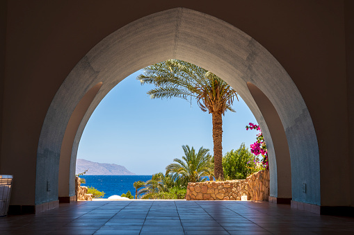Beautiful arched doorway in a building on the shores of the Red Sea at morning in the resort town of Sharm El Sheikh, Egypt, Africa. Blue sea, green palm tree and arch on the beach