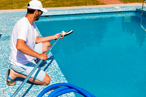 Mature man in flip flops cleaning the swimming pool with a vacuum cleaner.  Man working as a cleaner of the swimming pool, he standing with special equipment for cleaning at poolside and working