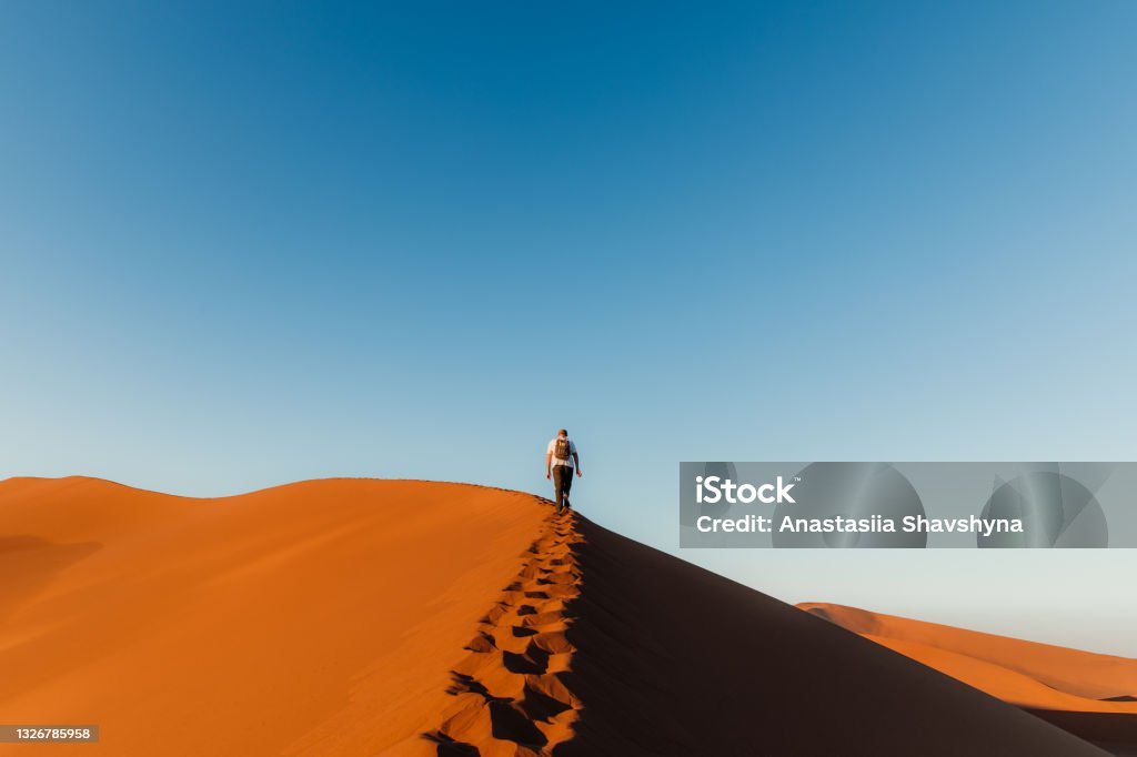 Man traveler enjoying the scenic sunrise from top of the dune at Sossuvlei, Namibia Man backpacker walking to the top of the sand dune looking at the picturesque bright sunrise at the desert in Namib-Naukluft National Park Individuality Stock Photo