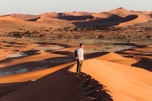 Young man tourist with backpack getting to the top of the dune looking at the bright sunny sunrise above the desert landscape at Namib-Naukluft-National Park