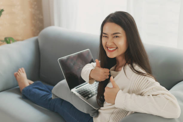 young happy asian wearing casual clothes using laptop computer relax sitting on couch sofa at home. asian employee with success expression showing thumps up looking at camera with smiley face - thumps up imagens e fotografias de stock
