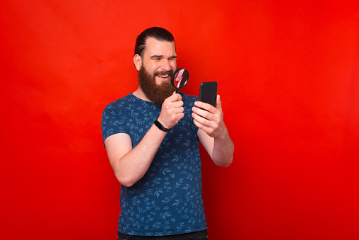 Photo of bearded man looking with expression trough magnifying glass at mobile phone, over red background