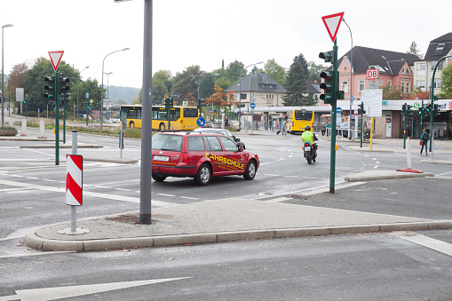 Driving school car and motorcycle are crossing intersection in front of station Essen Werden. In background a some people and a bus