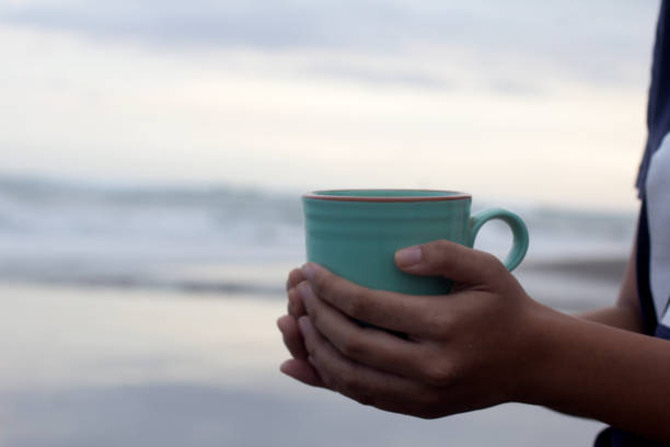 personne tenant une tasse de café ou de thé dans les mains sur le fond de vacances à la plage. - at the beach photos et images de collection