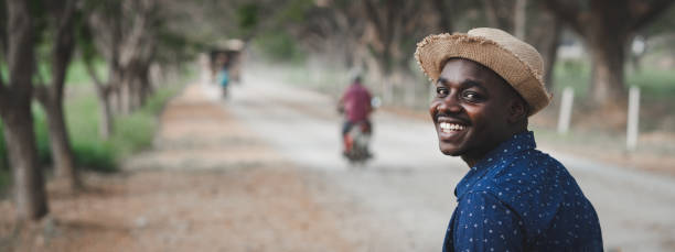 African man is wearing a hat and smiling while standing beside a rural road stock photo