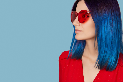 Happy young woman in red dress and sunglasses with blue hair on blue background. Fashion shopping sale concept