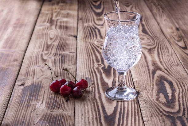 l'acqua frizzante fresca viene versata in un bicchiere, le bacche di ciliegio giacciono accanto ad essa su uno sfondo di legno. messa a fuoco selettiva - soft cider foto e immagini stock