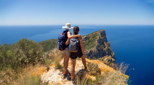 Two women hikers on mountain summit overlooking the mediterranean sea Two women hikers on mountain summit overlooking the mediterranean sea. Outdoor adventure day trek with friends. Aspiration, exploration and inspiring lifestyle concept. Panoramic from a mountain peak. majorca stock pictures, royalty-free photos & images