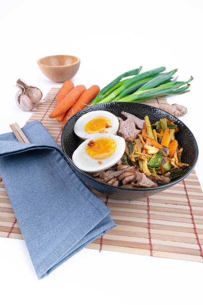 plan vertical et large en studio d’un bol noir de nouilles ramen avec des œufs durs, des légumes et de la dinde se rencontrent, prêt à être versé avec un bouillon bouillant. baguettes en bois. nourriture asiatique fraîchement préparée. - asian cuisine blue japanese culture still life photos et images de collection