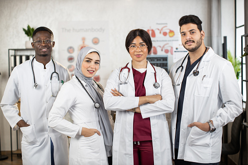 Team of female and male multiracial doctors in white lab coats keeping hands crossed or in the pockets while standing together at medical center. Concept of people, medicine and teamwork.