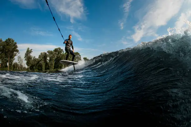 young athletic man in wetsuit vigorously riding on the wave with foilboard on background of sky and trees