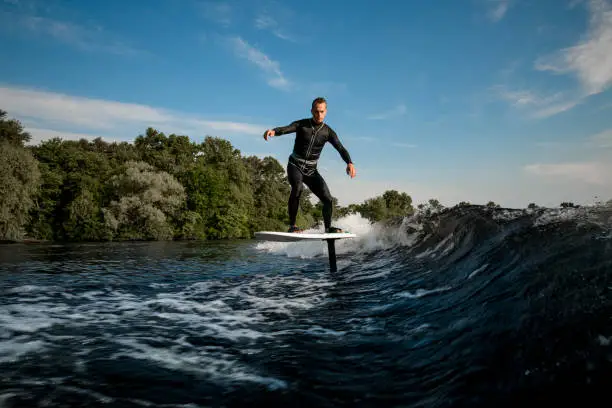 healthy athletic man in black wetsuit balancing on hydrofoil foilboard on a wave on a sunny day