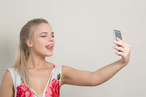 Playful blonde girl showing her tongue and making selfie on mobile phone