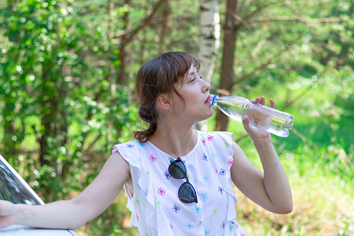 Beautiful young woman drinks water on a summer sunny day against a background of green foliage. Selective focus