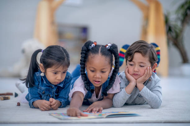 Three multi ethnic preschool children reading a book Three adorable multi ethnic preschool children are lying down next to each other on their classroom floor. They have a book open in front of them. They are focused on reading the book together. montessori stock pictures, royalty-free photos & images