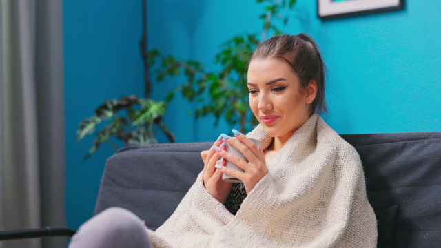 Happy young woman relaxing at home wrapped in a warm blanket on the sofa surfing the internet on her laptop computer with a smile of pleasure