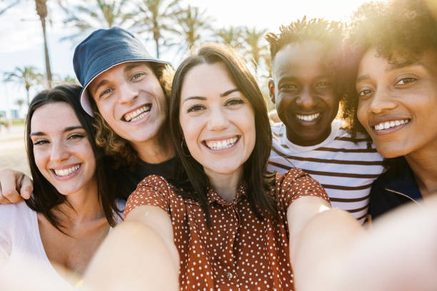 istock Group of happy multiracial people taking a selfie with mobile phone outdoors with back sunlight - Multiethnic group of friends in summer clothes having fun on holidays in the beach with tree palms on the background 1326846882