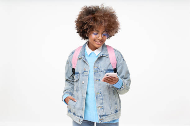 niña de la escuela africana con peinado afro y mochila sosteniendo el teléfono inteligente con una mano, enviando mensajes de texto a un amigo, usando la aplicación de redes sociales, aislada sobre fondo gris - vertical caucasian glasses red hair fotografías e imágenes de stock