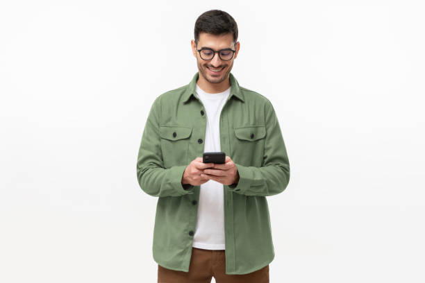 灰色の背景に孤立して立って、電話を見て緑のカジュアルシャツを着た若い男 - 隔離状態 ストックフォトと画像