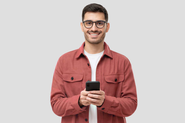 istock Young modern man holding smart phone in hands, looking at camera with smile, isolated on gray background 1326852410