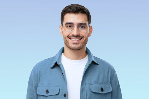 Headshot portrait of young it guy face wearing round glasses, looking at camera with smile, isolated on blue background