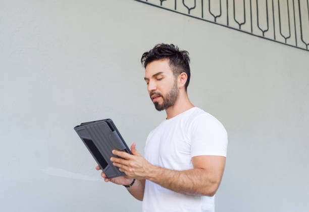 istock Handsome man standing in front of a gray wall scrolling and typing on electronic tablet device. 1326857549