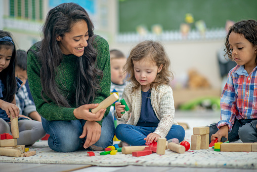 Preschool age children are seated on their classroom floor playing with wooden blocks. Their female teacher is seated next to them and helping one of the children with her blocks.