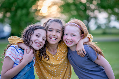 Three adorable girls around ten years old are standing close to each other outdoors. They are embracing each other and smiling directly at the camera.