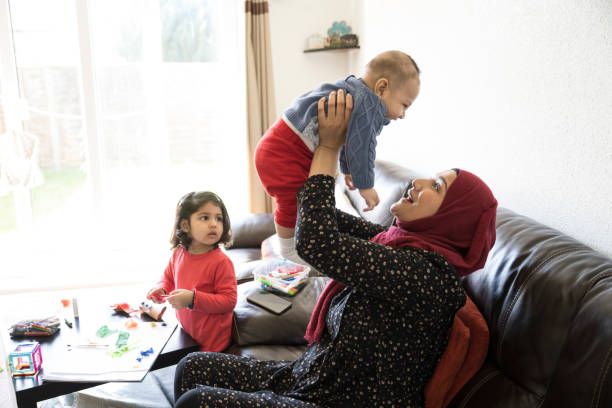 Early 30s British Asian mother playing with baby son at home Side view of woman in headscarf and tunic sitting on sofa in living room lifting her laughing 5 month old boy while toddler daughter works on craft project. hijab photos stock pictures, royalty-free photos & images