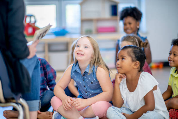 Caucasian girl with Down syndrome in a classroom A multi ethnic group of elementary school children are seated together on a classroom floor. They are looking at their teacher and listening intently as their teacher reads them a story from a book. special education stock pictures, royalty-free photos & images