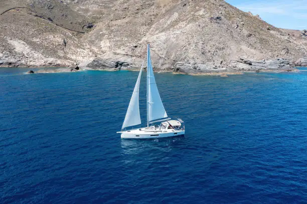 Summer vacations in Cyclades islands. Greece. Sailboat with open white sails cruising calm rippled sea, aerial drone view. Sunny day. Sailing in Aegean sea