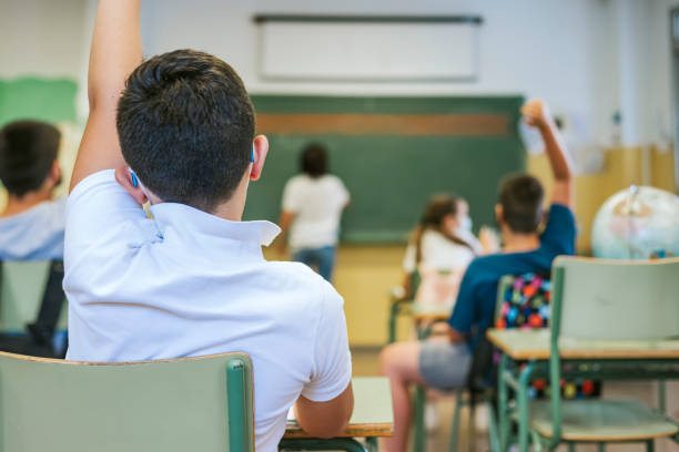A boy and his classmates raise their hands as the teacher writes on the blackboard. Back to school concept stock photo