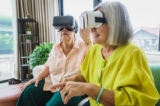 Asian senior woman friends enjoy using virtual reality glasses together in living room at home