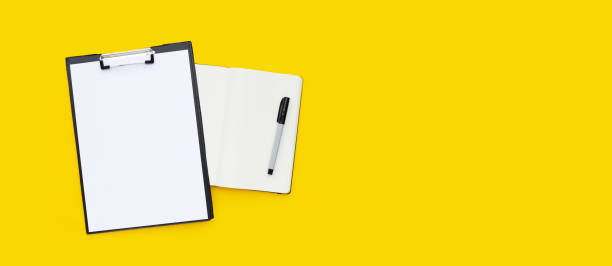istock Blank a4 paper in  black clipboard with notebook and pen on yellow background. 1326903459