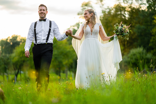 beautiful happy smiling romantic wedding couple walking together hand in hand through summer meadow at sunset, bride with bridal flowers looking at her husband