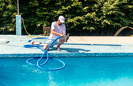 Mature man in flip flops cleaning the swimming pool with a net.  Man working as a cleaner of the swimming pool, he standing with special equipment for cleaning at poolside and working