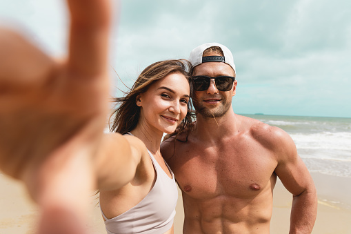 Happy young sport couple taking a selfie by smartphone at the seaside. Healthy man and woman friend take photograph together after workout at the beach