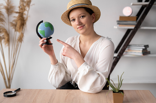 tourist girl shows on the globe a place for travel. The traveler in the office with the world in his hands. A young woman in a straw hat organizes tours at the agency. Travel concept.